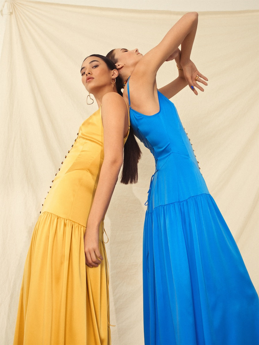Two sustainable, handmade maxi dresses in gold and bright blue with tiered skirts, worn by models in a minimalist setting.