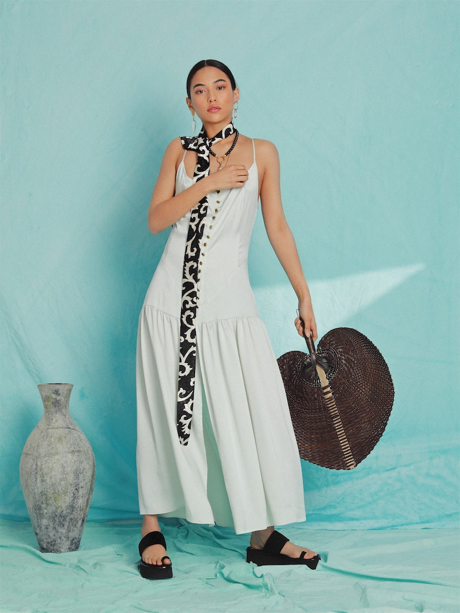 White sustainable maxi dress with thin straps and a tiered skirt, styled with black sandals and a patterned black and white.