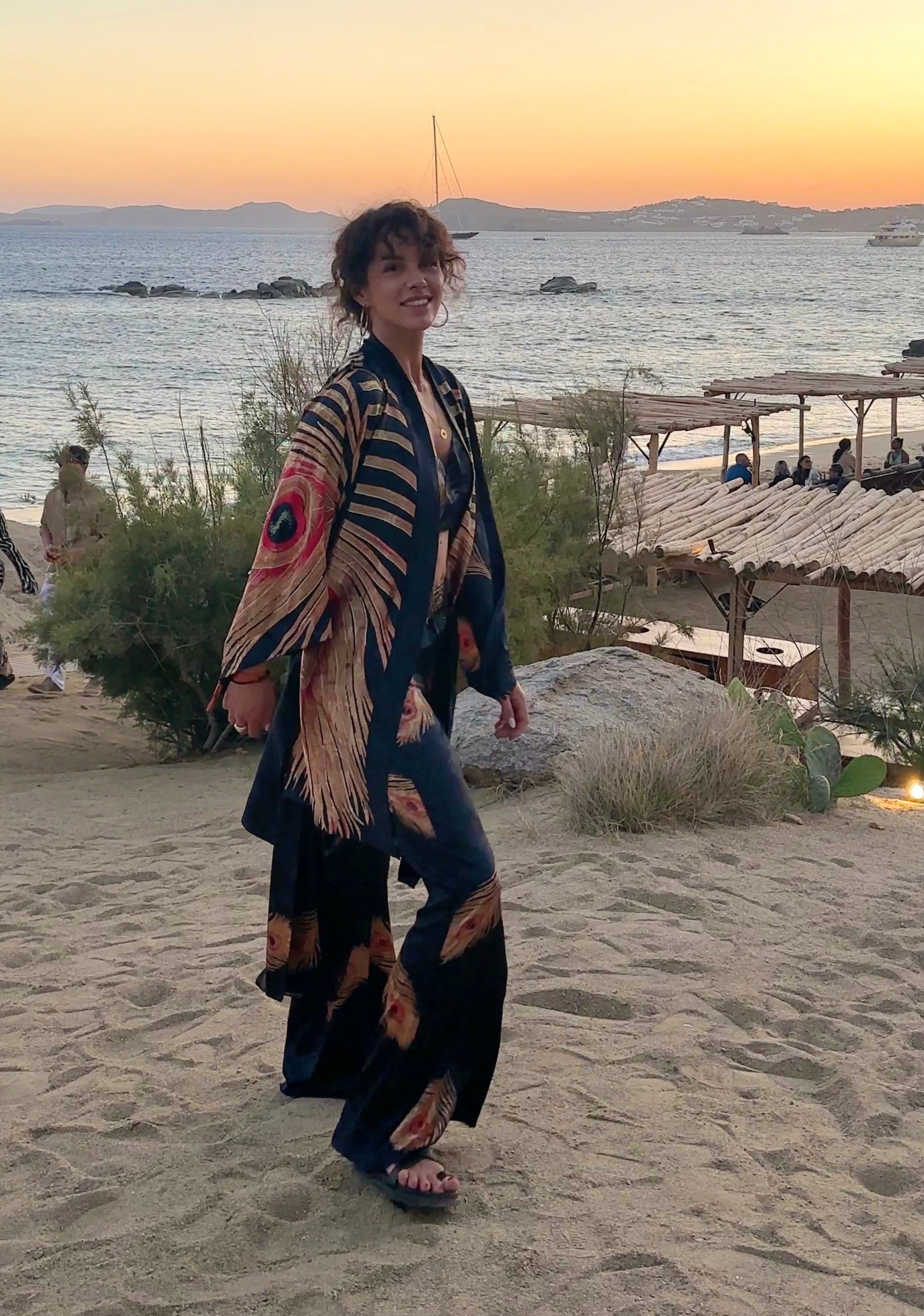 Woman dressed in black silk set walking on a sandy beach at sunset in Mykonos